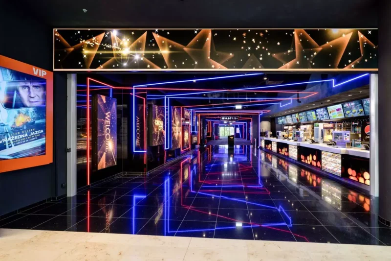 Svetlý vchod do kina s neónovo modrými a červenými svetlami, lesklou reflexnou podlahou a stánkom s občerstvením vpravo. Steny lemujú plagáty, ktoré vytvárajú živú a prívetivú atmosféru.