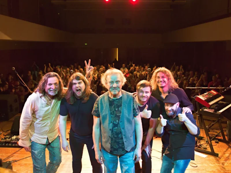 Šesť usmiatych členov kapely stojí na pódiu a pózuje na skupinovej fotografii s jásajúcim publikom v pozadí. Scéna je radostná a energická.