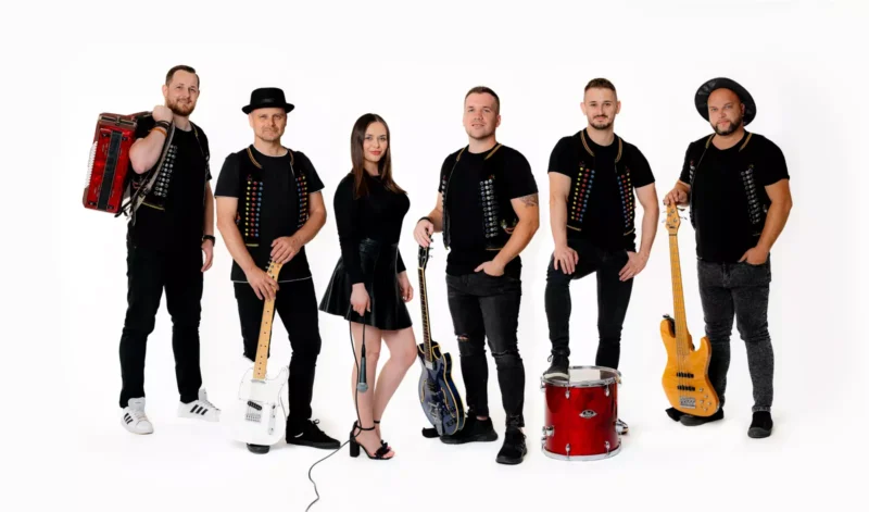 Skupina šiestich hudobníkov pózuje na bielom pozadí. Držia nástroje ako akordeóny, gitary a bubon. Všetci majú na sebe čierne košele a vyzerajú sebavedomo.
