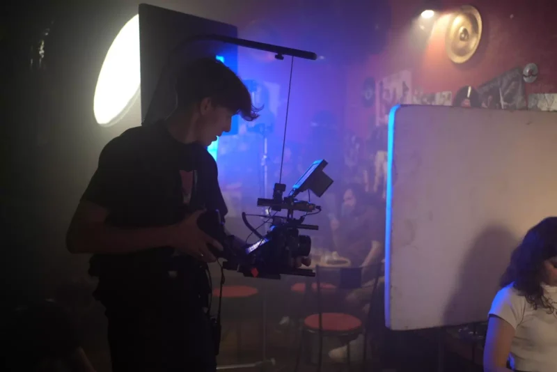 Filmár drží kameru v slabo osvetlenej miestnosti s červenými stenami a hudobnými plagátmi. Jemné osvetlenie a uvoľnená atmosféra naznačujú neformálne prostredie natáčania.