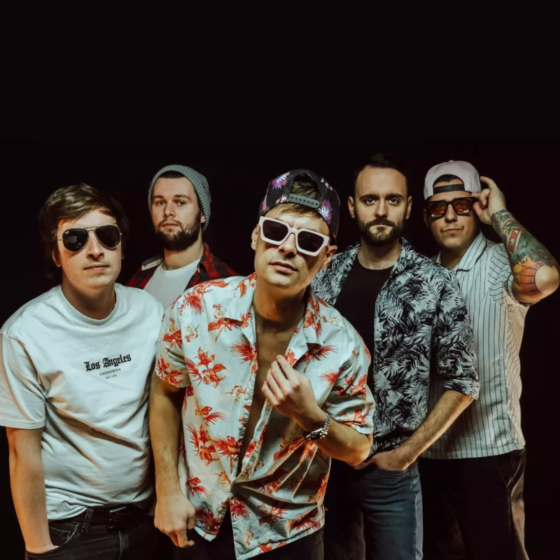 Skupina piatich mužov stojí sebavedomo v rade, oblečení v tropických košeliach a slnečných okuliaroch. Atmosféra je neformálna a živá, s atmosférou pripomínajúcou kapelu.