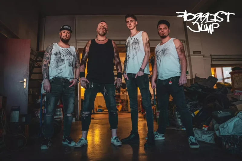 Štyria muži so špinavými tielkami a tetovaniami stoja sebavedomo v slabo osvetlenej garáži. Ich výrazy vyjadrujú húževnatosť a jednotu. Na stene je graffiti.