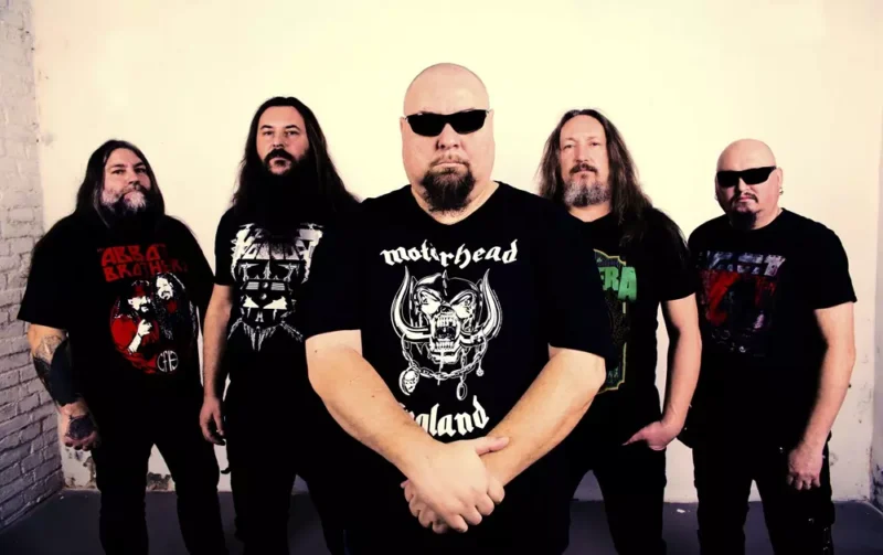 Päť mužov s vážnymi výrazmi v tvári stojí pri bielej stene a majú na sebe čierne tričká s potlačou. Ústredná postava má na sebe slnečné okuliare, ktoré vytvárajú odvážnu a intenzívnu atmosféru.