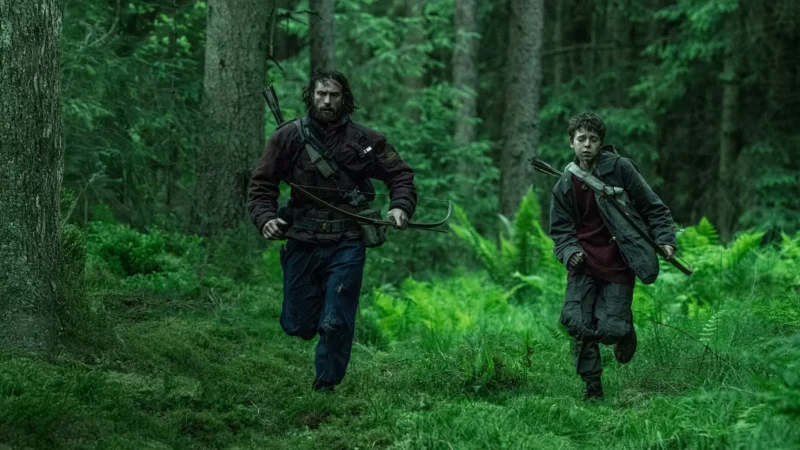 Dvaja ľudia bežia hustým, bujným lesom. Jeden drží luk, obaja vyzerajú sústredene a odhodlane, vyjadrujúc naliehavosť a napätie.