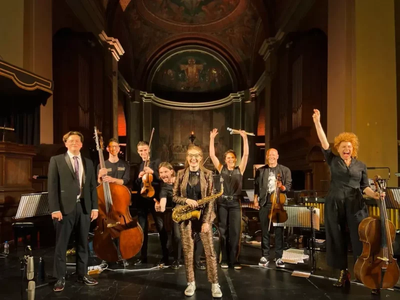 Radostná skupina ôsmich hudobníkov stojí na slabo osvetlenom pódiu vo veľkej sále, drží nástroje ako violončelo a saxofón a oslavuje so zdvihnutými rukami.