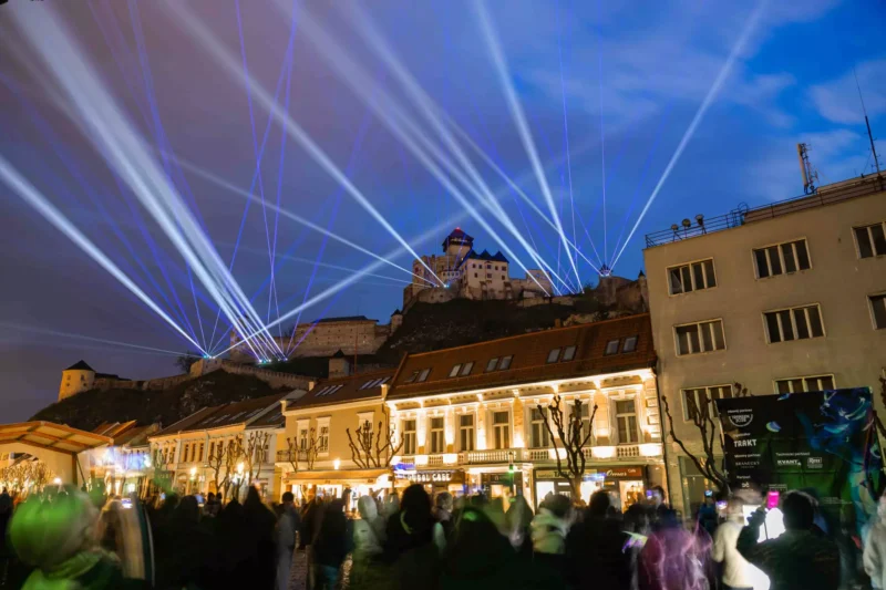 Mestská ulica plná ľudí v noci, osvetlená žiarivými modrými laserovými lúčmi vychádzajúcimi z trenšianskeho hradu na kopci pod zamračenou oblohou. Slávnostná atmosféra.