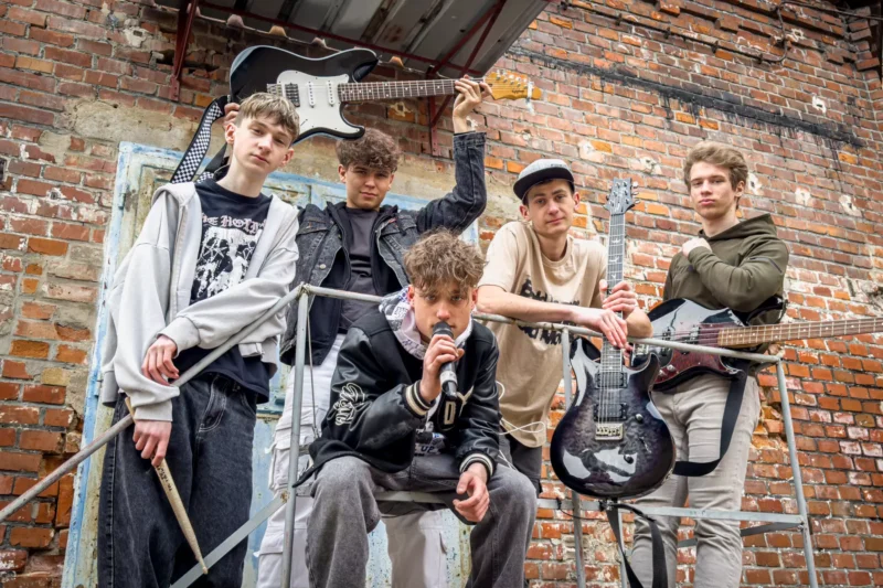 Piati mladí muži pózujú pri zvetranej tehlovej stene, z ktorých vyžaruje atmosféra rebelskej rockovej kapely. Jeden drží mikrofón, ďalší gitary a paličky.