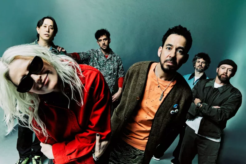 Rozmanitá skupina šiestich ľudí sa nakláňa k fotoaparátu a vyžaruje z nej ležérna a hravá atmosféra. V popredí sa usmieva žena v červenom a slnečných okuliaroch.