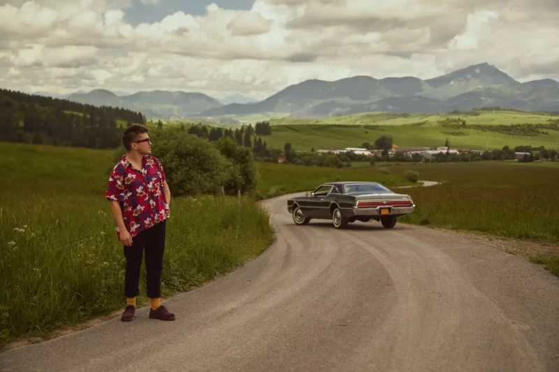 Muž v kvetinovej košeli stojí na kľukatej vidieckej ceste a pozerá sa na klasické auto pod zamračenou oblohou s horami v pozadí. Pokojná retro atmosféra.