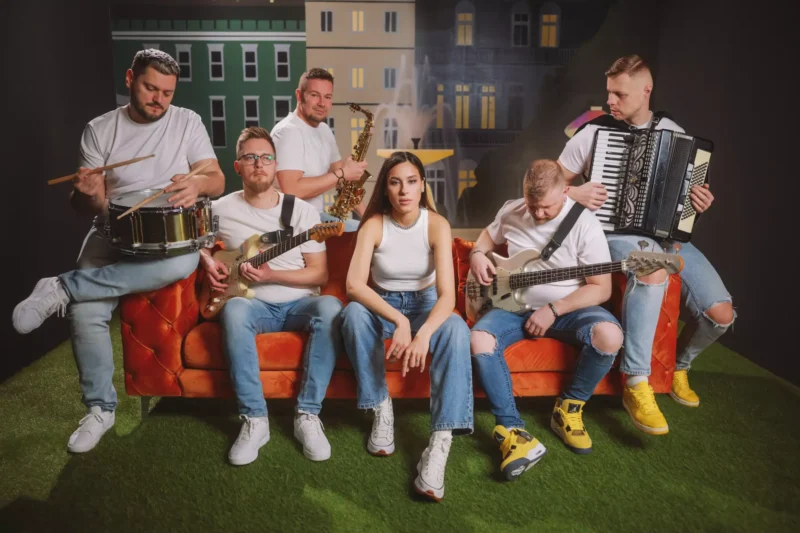 Skupina šiestich hudobníkov na oranžovom gauči v bielych košeliach a džínsoch s pozadím panorámy mesta. Medzi nástroje patria bicie, gitara, saxofón, basgitara a akordeón. Nálada je uvoľnená a spolupracujúca.