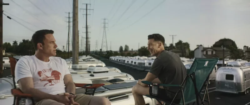 Dvaja muži sedia na kempingových stoličkách na karavane, jeden má na sebe bielu košeľu, druhý sivú. Rozprávajú sa pri západe slnka, na pozadí radu obytných áut a elektrického vedenia, čo navodzuje uvoľnenú, úprimnú atmosféru.