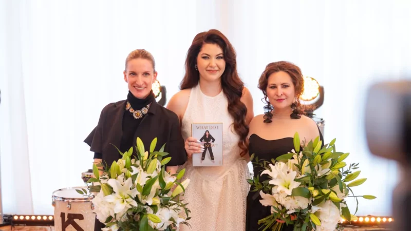 Tri ženy stoja s úsmevom, jedna v bielych šatách drží knihu s názvom „Čo viem?“. Ostatné v čiernych outfitoch držia veľké kytice bielych ľalií.