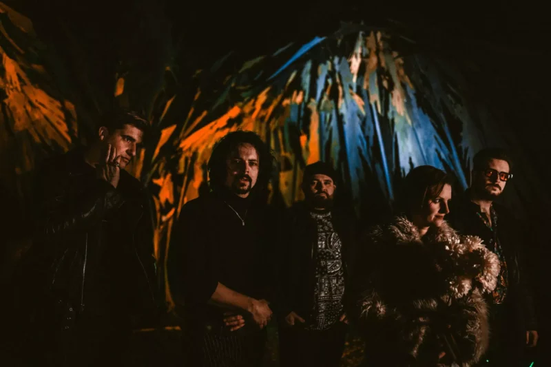 Päť ľudí stojí v tlmenom prostredí s farebným, rozmazaným abstraktným umením ako kulisou. Ich výrazy sú vážne a vytvárajú tajomnú, náladovú atmosféru.