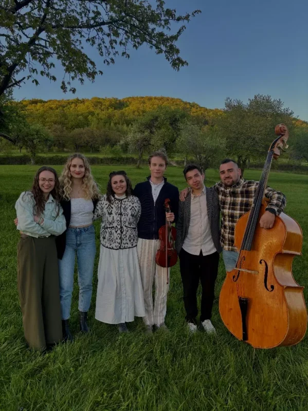 Skupina šiestich ľudí stojí tesne pri sebe na trávnatej lúke pri západe slnka a vrúcne sa usmievajú. Jeden drží husle a druhý kontrabas, čo evokuje hudobné ladenie.