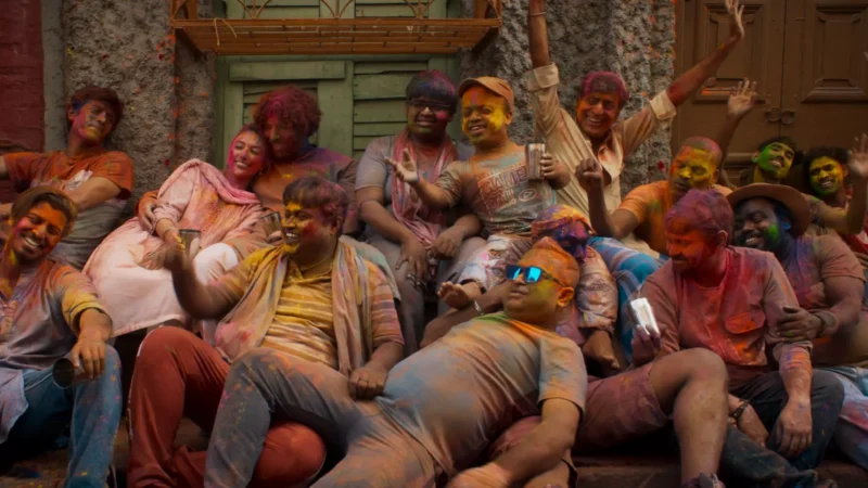 Radostná skupina oslavuje Holi, zahalená v žiarivých farbách, sediaca pri rustikálnom múre. Úsmevy a smiech vytvárajú slávnostnú a bezstarostnú atmosféru.