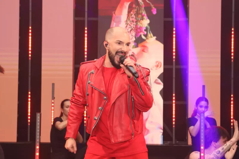 Účinkujúci v žiarivo červenom saku vášnivo spieva na pódiu s mikrofónom v ruke. Živú atmosféru umocňujú tanečníci v pozadí a farebné svetlá pódia.