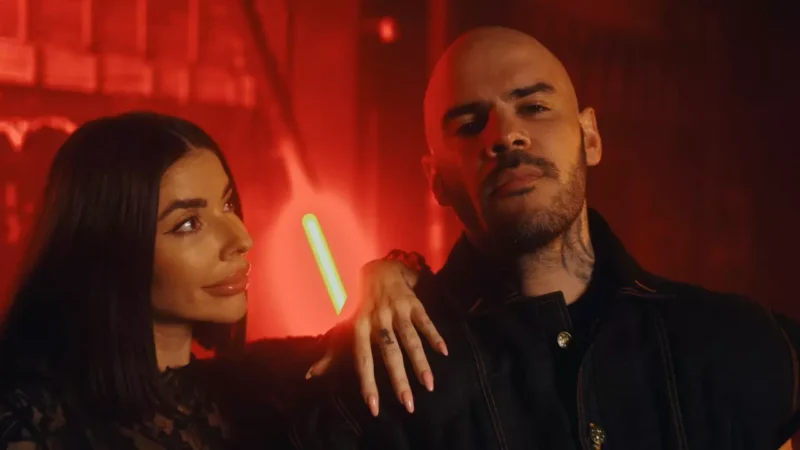 Žena s dlhými tmavými vlasmi sa pozerá na plešatého muža s tetovaniami v slabo osvetlenej červenej miestnosti. Atmosféra je tajomná a intenzívna.