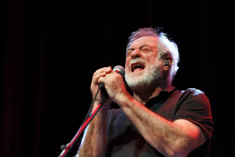 Starší muž vášnivo spieva do mikrofónu na pódiu so zatvorenými očami a intenzívnym výrazom, pod tmavým pozadím a v svetle reflektorov.