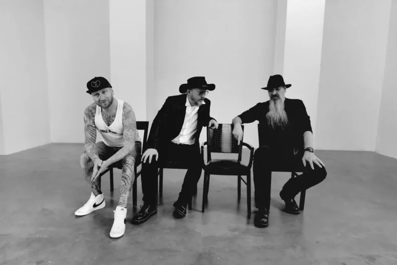 Traja muži sedia na stoličkách v minimalistickej miestnosti. Muž naľavo má tetovania a čiapku a ďalší dvaja majú na sebe čierne klobúky a oblečenie.