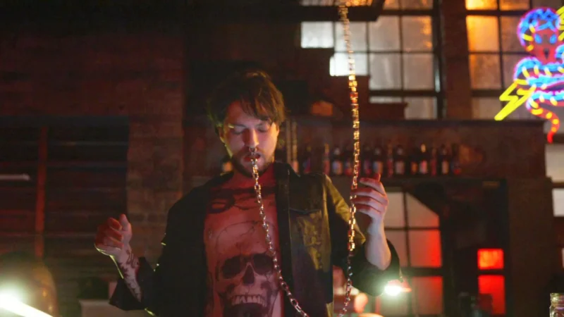 Muž v čiernej bunde a košeli s lebkou si v slabo osvetlenom bare s neónovými svetlami a fľašami v pozadí vyťahuje retiazku z úst.