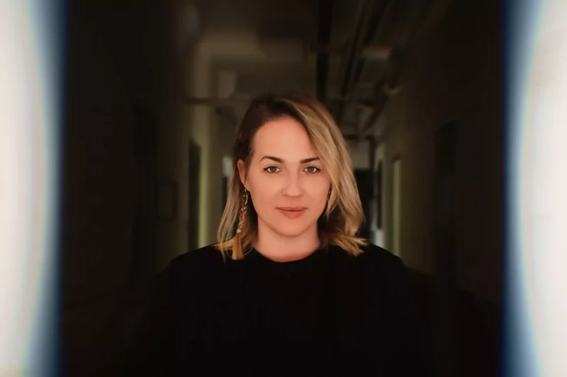 Portrét ženy s blond vlasmi a náušnicami, stojacej v slabo osvetlenej chodbe. Pozadie je rozmazané, čo vyjadruje pocit sústredenia a pokoja.