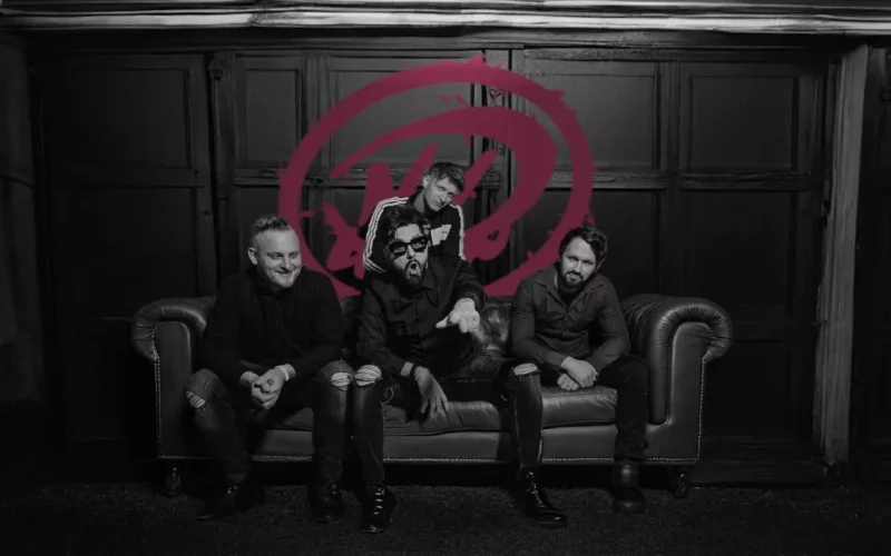 Štyria ľudia sedia na gauči v čiernobielej miestnosti. Jeden energicky dvíha ruku. Na stene za nimi je červený, abstraktný kruh s čiarou.