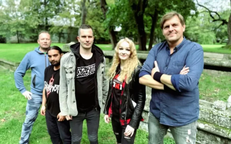 Skupina piatich ľudí stojí vonku v parku a ležérne sa usmieva. Svieža zelená kulisa so stromami a rustikálnym plotom vytvára uvoľnenú a priateľskú atmosféru.