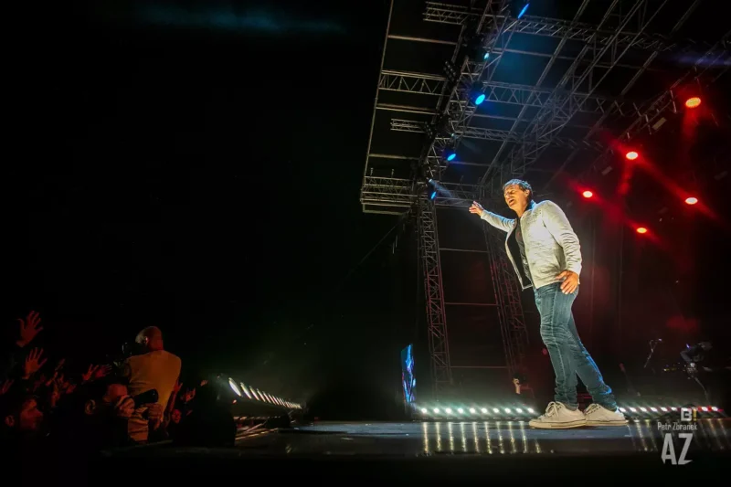 Umelec na osvetlenom pódiu ukazuje na jásajúci dav a vytvára tak energickú a živú atmosféru. Svetlá pódia svietia červenou a modrou farbou.