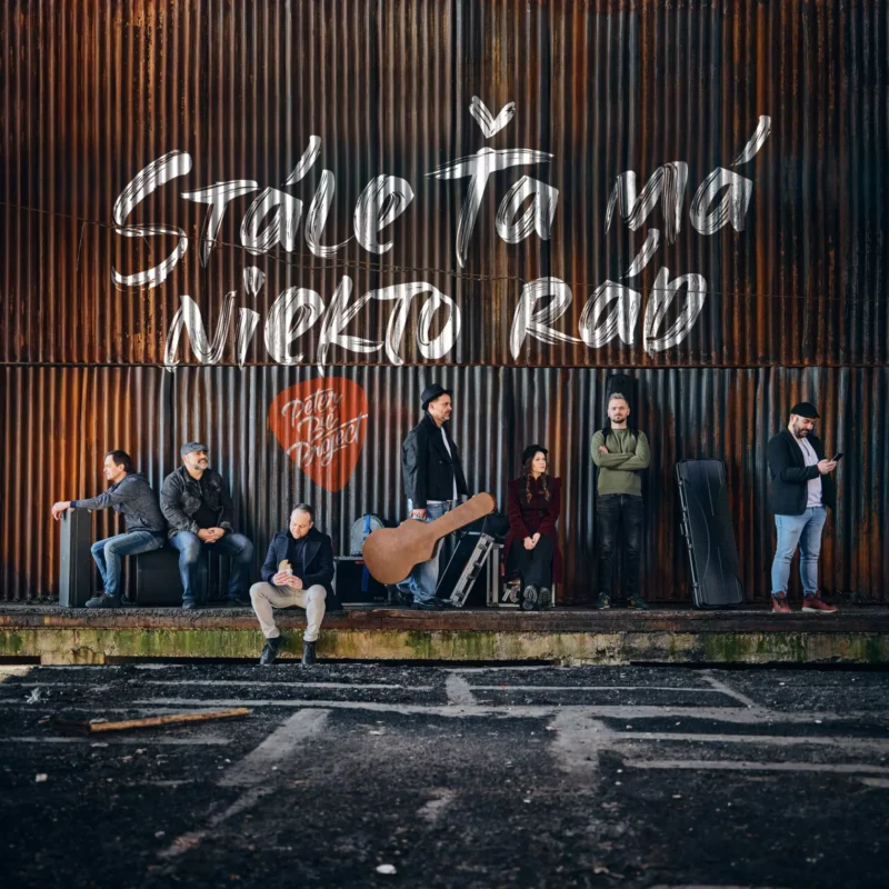 Sedem ľudí stojí a sedí pri veľkej, hrdzavej vlnitej kovovej stene s nápisom „Stále Ťa má Niekto Rád.“ Nálada je ležérna a zamyslená.