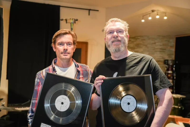 Dvaja muži sa usmievajú a držia zarámované vinylové platne v príjemne osvetlenej miestnosti. Jeden má na sebe kockovanú košeľu a druhý čierne tričko, čo vyjadruje pocit úspechu.