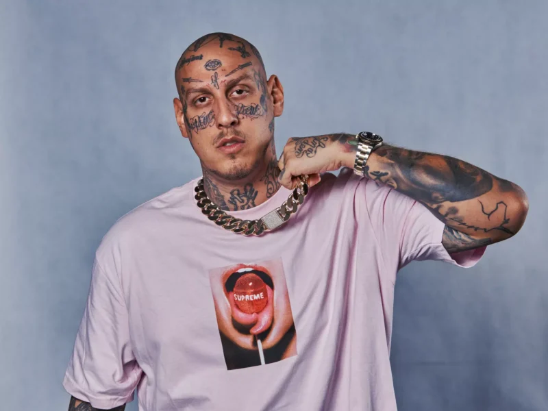 Muž s tetovaniami na tvári a ruke má na sebe svetloružové tričko s perami a lízankou. Drží zlatú retiazku a pozerá sa priamo do kamery. Sebavedomý tón.