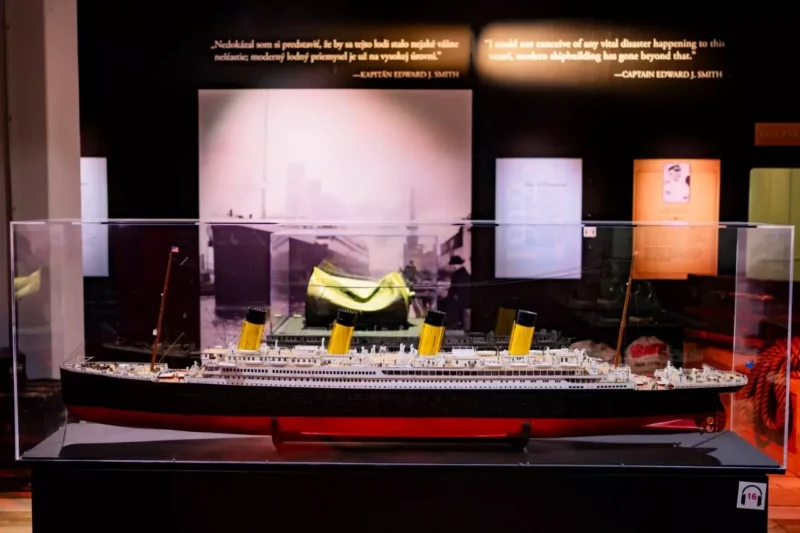Model lode RMS Titanic zaliaty v skle s ikonickými žltými lievikmi. Pozadie obsahuje historický text a obrázky, ktoré vytvárajú nostalgickú atmosféru.