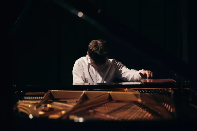 Klavirista v bielej košeli sa skláňa nad klavírom, ruku má položenú na notovom zábere. Tlmené osvetlenie vytvára sústredenú, kontemplatívnu atmosféru.