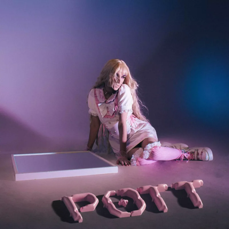 Osoba v ružovo-bielom kostýme s dlhými blond vlasmi pózuje na podlahe v slabo osvetlenej miestnosti. Na zemi sú napísané ružové písmená s nápisom „POTT“.