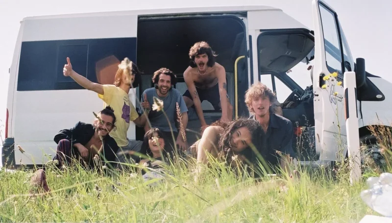 Radostná skupinka priateľov energicky pózuje pred bielou dodávkou zaparkovanou na bujnej lúke, so svietiacim slnkom a posiatymi divými kvetmi okolo.
