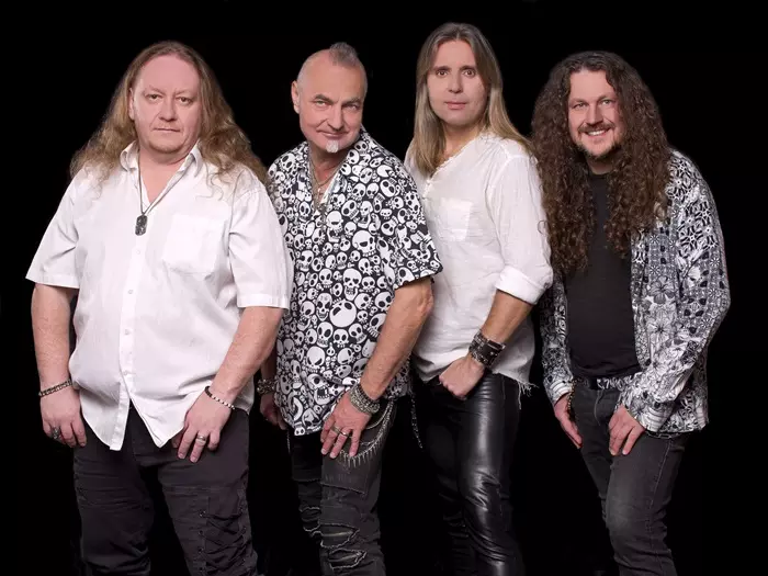 Štyria muži v rockovej kapele pózujú na čiernom pozadí. Majú na sebe vzorované košele a kožené nohavice, z ktorých vyžaruje sebavedomie a kamarátstvo.