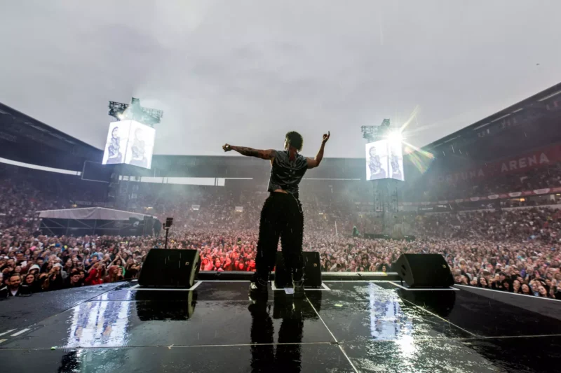 Účinkujúci na pódiu čelí jasajúcemu davu vo veľkej vonkajšej aréne. Scéna je živá, dážď a dramatické osvetlenie vytvárajú energickú atmosféru.