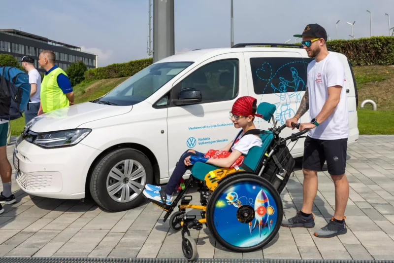 Muž tlačí dieťa na pestrofarebnom invalidnom vozíku s raketovými vzormi vedľa bielej dodávky. Scéna vyvoláva podporujúcu a radostnú atmosféru.
