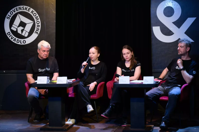 Na pódiu v Slovenskom komornom divadle sedia štyria ľudia a zúčastňujú sa panelovej diskusie. Majú na sebe čierne tričká s textom a pôsobia sústredene a pozorne.