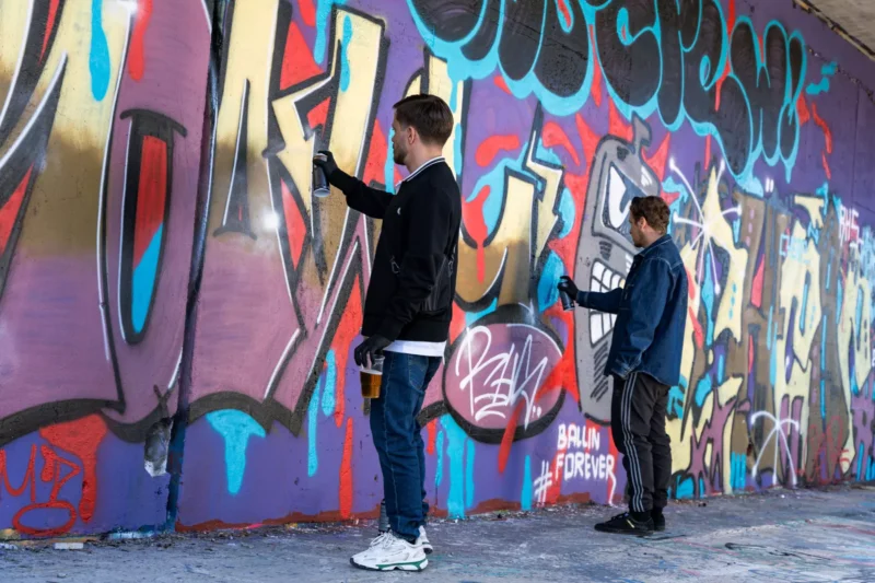 Dvaja ľudia sprejujú žiarivú graffiti stenu výraznými farbami ako červená, modrá a zlatá. Majú na sebe ležérne oblečenie a čierne rukavice a sústredia sa na svoje umenie.