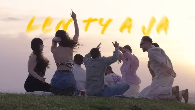 Skupina ľudí sedí na trávnatom kopci pri západe slnka, otočení chrbtom k fotoaparátu. Vyzerajú radostne, dvíhajú ruky a energicky spolu komunikujú.