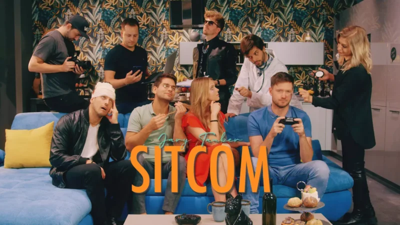 Skupina ľudí vo farebnej miestnosti; niektorí s kamerami, iní hrajú. Jedna osoba drží herný ovládač. Nápis „Sitcom“. Veselá scéna.