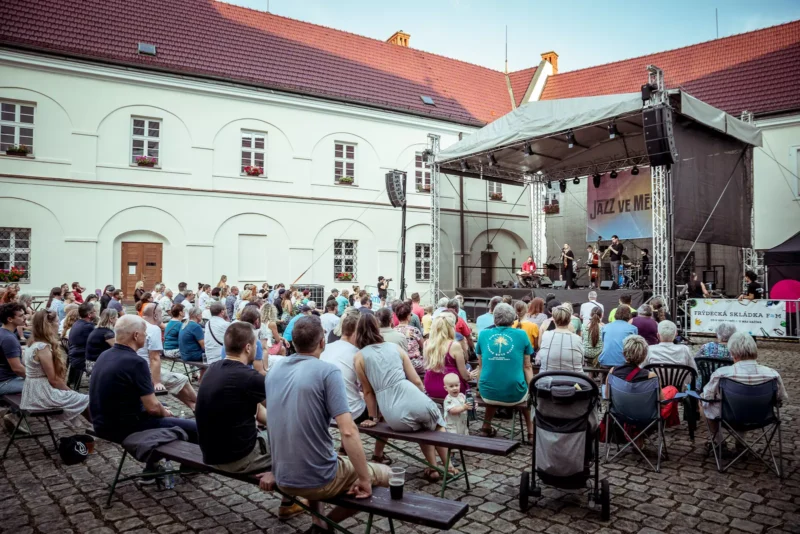 Vonkajší jazzový koncert na nádvorí. Publikum sedí na lavičkách oproti pódiu, kde vystupujú hudobníci. Atmosféra je živá a uvoľnená.