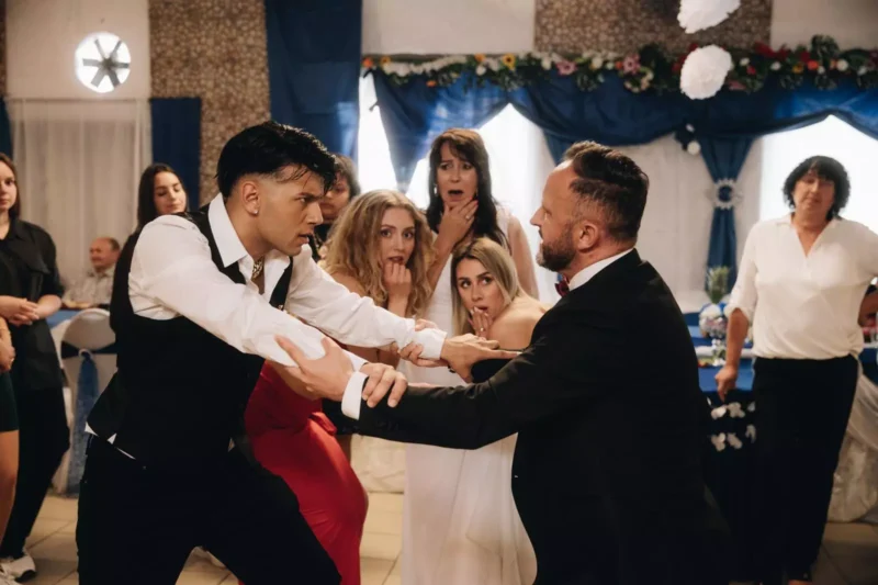 Napätá scéna na svadobnej hostine, kde sa dvaja muži v oblekoch stretávajú a držia sa za ruky. Prizerajúci sa v spoločenských šatách vyzerajú šokovane a znepokojene.