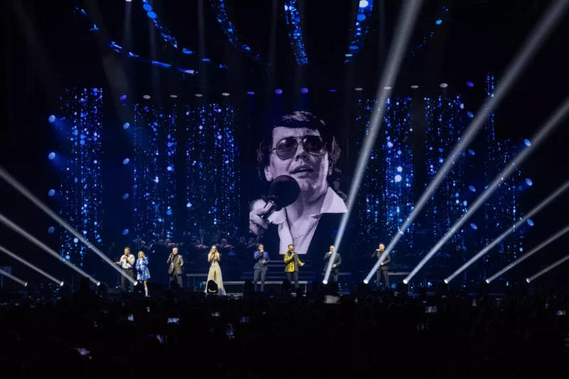 Skupina účinkujúcich spieva na živom pódiu s modrými svetelnými efektmi a reflektormi. Na veľkej obrazovke sa premieta čiernobiely obraz muža s mikrofónom.