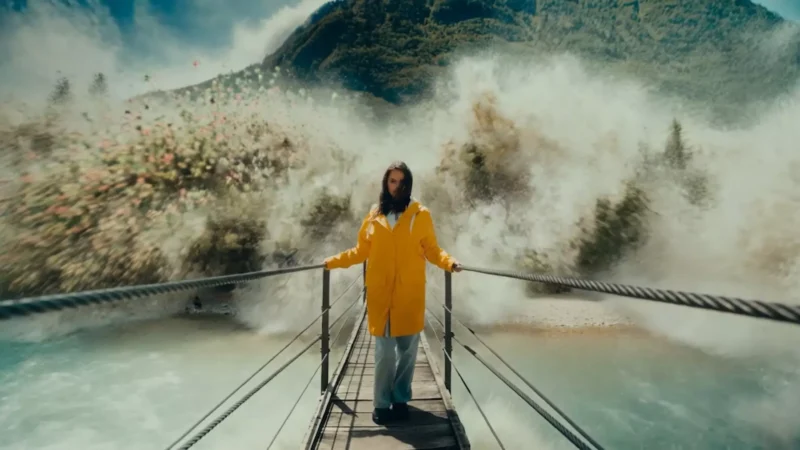 Žena v žltom pršiplášti stojí na úzkom visutom moste nad dynamickou, hmlistou riekou. Za ňou kontrastuje žiarivá explózia kvetov so zelenou horou pod zamračenou oblohou.