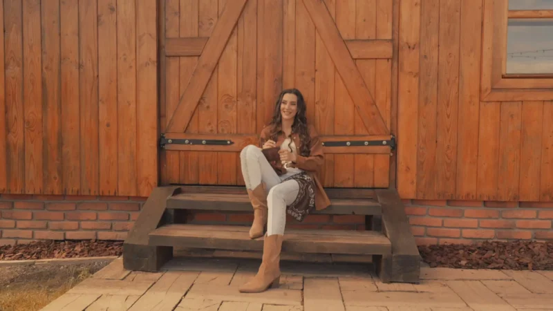 Usmievavá žena sedí na drevených schodoch pred rustikálnymi dverami stodoly. Má na sebe hnedú bundu, biele nohavice a hnedé čižmy a v ruke drží nápoj. Scéna je teplá a útulná.