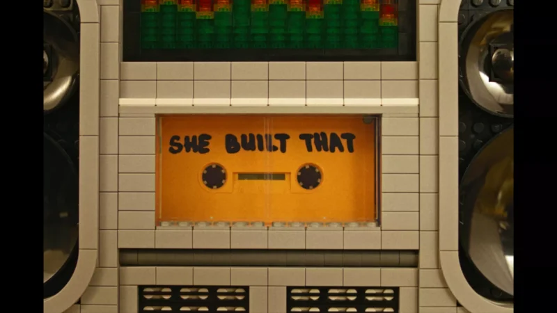 Detailný záber na boombox z lega s oranžovou kazetou a nápisom „She Built That“ (Ona to postavila). Tón je nostalgický a kreatívny.
