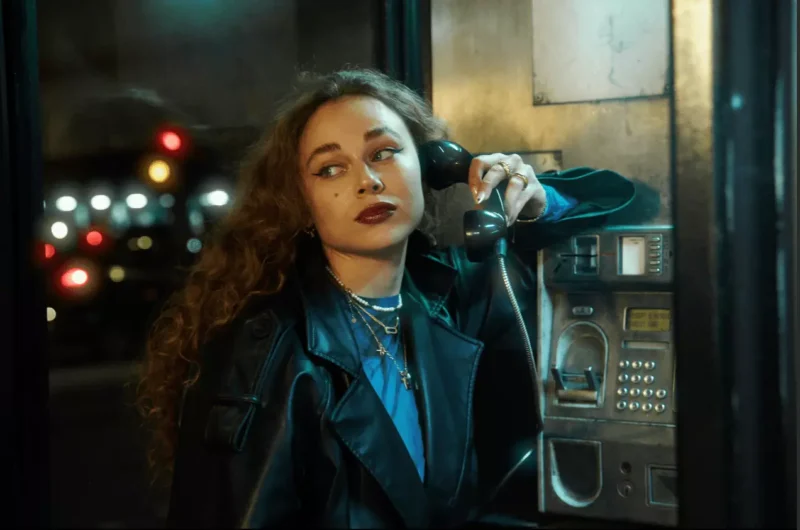 Žena v koženej bunde používa v noci telefónnu búdku. Zamyslene sa pozerá inam. Svetlá mesta vytvárajú jemný bokeh efekt a dodávajú nostalgickú atmosféru.
