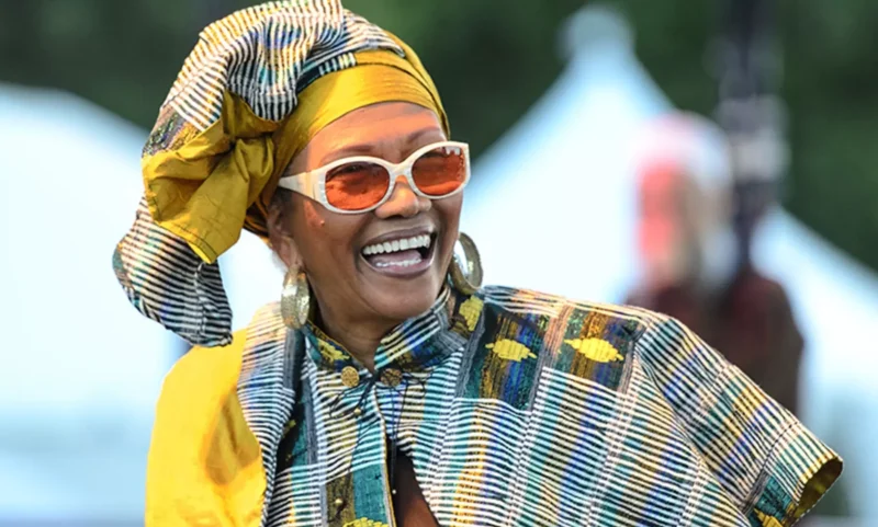 Žena sa radostne usmieva v žiarivom, vzorovanom oblečení a s pokrývkou hlavy v žlto-modrých tónoch. Má biele okuliare a vyžaruje z nej šťastie a energiu.